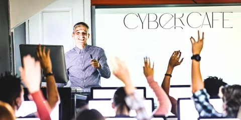 A class takes place in a room with several computers. Many hands are raised. A man with glasses stands at the front of the room, prepared to answer. 