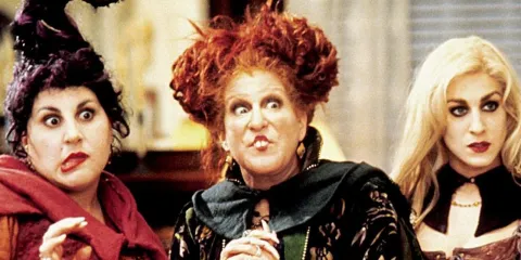 The three witches make silly expressions. One has flame-red hair, one pale blonde, and the other black curls obscured by a tall pointy hat. They all wear crimson lipstick. 