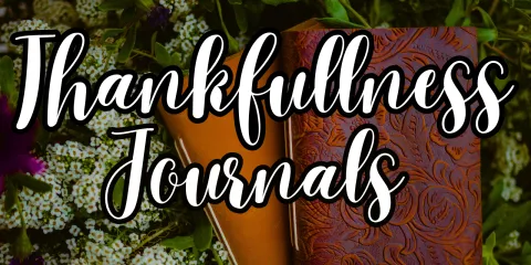 The words "Thankfulness Journals" is positioned, slanted, over a dark image. In this image, there are two journals - one with various plant shapes embellished on the cover and the other plain - resting on white flowers and green leaves.