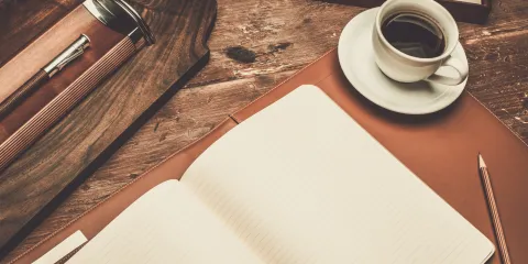 A blank notebook is spread over a table. Beside it is a brown pencil and black coffee. 
