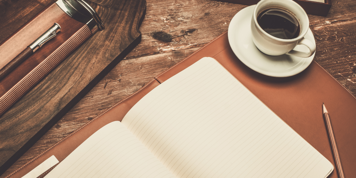 A blank notebook is spread over a table. Beside it is a brown pencil and black coffee. 